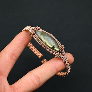 Può includere: Un bracciale a fascia realizzato con filo di rame avvolto, caratterizzato da una pietra preziosa di labradorite centrale a forma di marquise con iridescenza verde e oro. La lavorazione del metallo presenta motivi intrecciati.