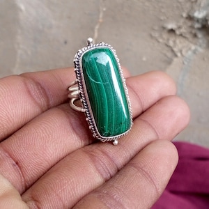 Könnte beinhalten: Ein Ring mit einem rechteckigen Malachit-Edelstein in einer Silberfassung. Der grüne Stein weist natürliche dunkle Streifenmuster auf. Die Silberfassung hat ein gedrehtes Kordelrand-Detail und ein mehrbändiges Ringschienen-Design.
