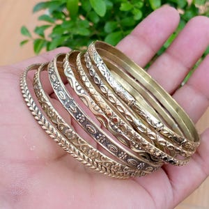 May include: A set of six gold-toned metal bangles featuring various embossed patterns including floral motifs, geometric lines, and textured borders. Each bracelet has a distinct design with a polished finish, stacked together to form a cohesive set.