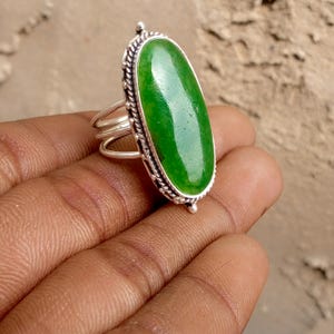 May include: A silver ring featuring a large, oval-shaped green gemstone. The gemstone is set in a detailed silver band with a rope-like border. The ring has a three-band design and is displayed on a hand.