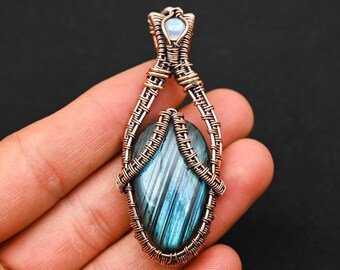 Peut inclure: Un pendentif composé d'une pierre de labradorite bleue en forme de goutte avec des inclusions linéaires sombres, enchâssée dans une armature complexe en fil de cuivre. Une petite pierre de lune ronde et irisée orne la bélière en cuivre.