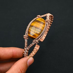 Könnte beinhalten: Kupferdrahtarmband mit einem großen, ovalen Tigeraugenstein. Der Stein hat braune und goldene Farbtöne. Die Drahtarbeit des Armbands ist aufwendig gewebt und schafft ein einzigartiges, handgefertigtes Design. Das Armband ist offen.