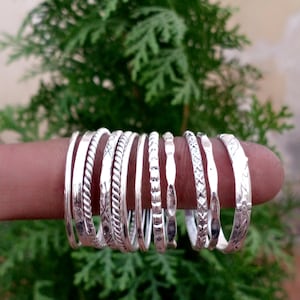 May include: A collection of ten silver-toned rings arranged in a row. The rings feature various textures and patterns, including smooth bands, twisted rope designs, etched geometric shapes, and small floral motifs, all displayed together on a finger.