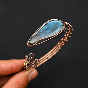 May include: A wire-wrapped cuff bracelet featuring a teardrop-shaped labradorite gemstone with blue iridescence. The band is constructed from copper wire with intricate woven patterns and a decorative terminal end with small gold-toned metal beads.