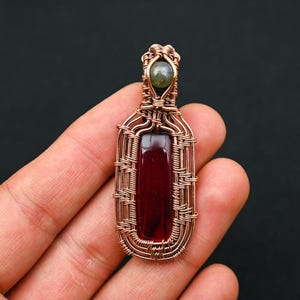 May include: A rectangular pendant featuring a deep red gemstone encased in intricate copper wire wrapping. A small, round, iridescent grey stone is set at the top of the wirework, which forms a decorative frame around the central red stone.