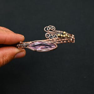Può includere: Bracciale in filo di rame con una gemma ovale viola chiaro. Il bracciale presenta volute decorative e piccole perline dorate. Il design è intricato e realizzato a mano.
