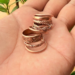 Puede incluir: Una pila de seis anillos de cobre con varios diseños, incluyendo una banda lisa, un patrón trenzado, un motivo floral y una textura geométrica. Los anillos son de color cobre metálico y varían en ancho, creando una apariencia superpuesta.