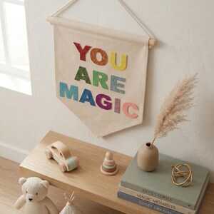 Può includere: Un banner di tela color crema con le parole "YOU ARE MAGIC" in lettere glitterate colorate è appeso a una parete. Sotto, una mensola di legno espone un'auto giocattolo, un giocattolo impilabile, un orsacchiotto, libri e oggetti decorativi.