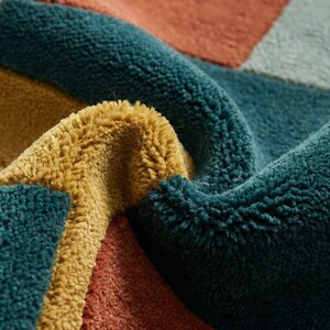 Modern abstract hand-tufted rug with geometric shapes and leaf pattern in teal, mustard, rust, and neutral tones placed in a stylish living room.