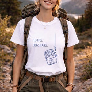May include: White t-shirt with the text "ZERO BITES. 100% SARDINES." and a blue illustration of a sardine can with fish. The shirt is short-sleeved and made of a soft material.