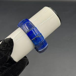 Könnte beinhalten: Ein lebendiges blaues Armband aus Lapislazuli. Das Armband besteht aus rechteckigen, polierten Steinen. Die Steine sind ein tiefes, sattes Blau mit Goldsprenkeln. Das Armband wird auf einem weißen zylindrischen Objekt präsentiert.
