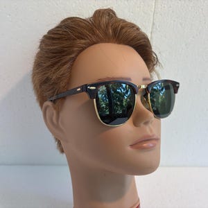 May include: Browline sunglasses featuring dark tortoiseshell upper frames and temples with gold-tone metal rims around the lower lenses. The lenses are dark green and reflective, showing a subtle outdoor reflection on their surface.