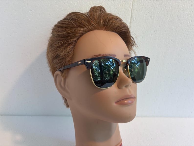 May include: Browline sunglasses featuring dark tortoiseshell upper frames and temples with gold-tone metal rims around the lower lenses. The lenses are dark green and reflective, showing a subtle outdoor reflection on their surface.