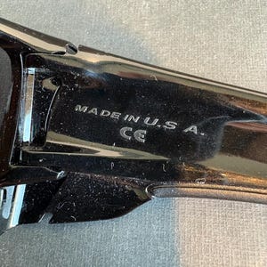 May include: Close-up of a black sunglasses frame with the text "MADE IN U.S.A." and the CE mark. The frame is glossy and reflects light, suggesting a polished finish. The image is taken against a neutral background.