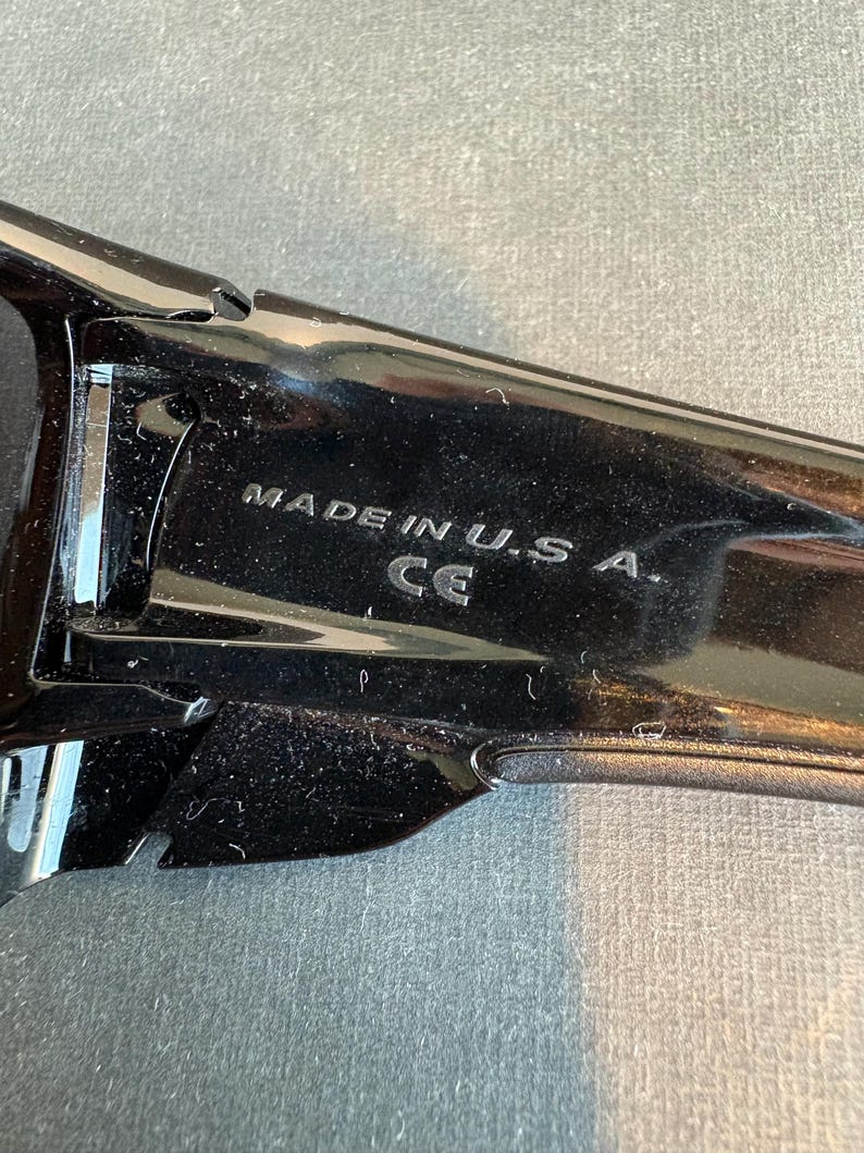 May include: Close-up of a black sunglasses frame with the text "MADE IN U.S.A." and the CE mark. The frame is glossy and reflects light, suggesting a polished finish. The image is taken against a neutral background.