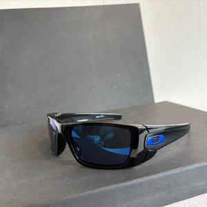 May include: Black sunglasses with blue mirrored lenses. The frames are black with a blue accent on the temple. The sunglasses are rectangular in shape and appear to be made of plastic.