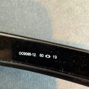 May include: Close-up of a black eyeglass frame with the text "OO9096-12 60 19" printed in white. The frame is glossy and appears to be made of plastic or a similar material. The image is well-lit, and the text is clear and easy to read.