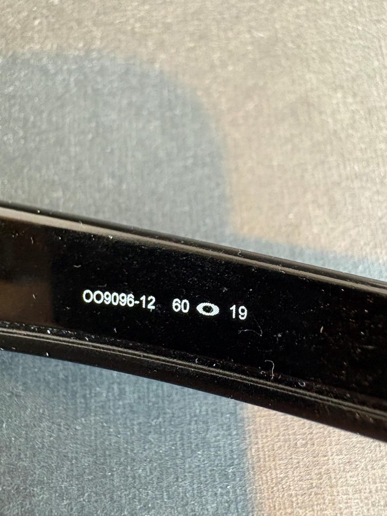 May include: Close-up of a black eyeglass frame with the text "OO9096-12 60 19" printed in white. The frame is glossy and appears to be made of plastic or a similar material. The image is well-lit, and the text is clear and easy to read.
