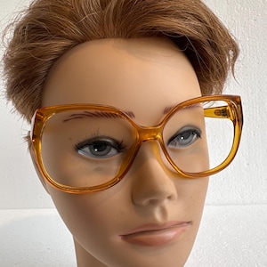 May include: Large, amber-colored eyeglasses with a thick, square frame. The glasses have a retro, oversized design, with a prominent browline and a slightly rounded lower edge. The lenses are clear, and the frames are a translucent, warm orange hue.