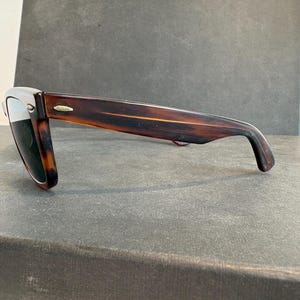 May include: Sunglasses with a tortoiseshell frame and dark lenses. The classic rectangular sunglasses have a subtle gold accent on the temples. The glasses are resting on a dark grey surface.