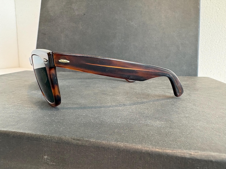May include: Sunglasses with a tortoiseshell frame and dark lenses. The classic rectangular sunglasses have a subtle gold accent on the temples. The glasses are resting on a dark grey surface.