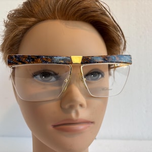 May include: Eyeglasses with a gold frame and a unique patterned top bar in shades of blue, brown, and gold. The lenses are clear, and the glasses have a rectangular shape. The brand name is printed on the lens.