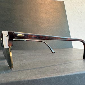 May include: Tortoiseshell-patterned sunglasses with a browline frame design. The frames feature dark brown and amber mottled acetate upper rims and temples, complemented by gold-tone metal lower rims and a small gold-tone accent on the temple hinge.