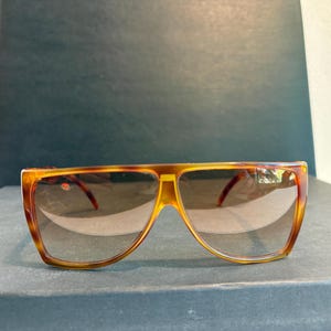 Puede incluir: Gafas de sol marrones y amarillas con estampado de carey. Las gafas tienen una montura ancha y rectangular y lentes tintadas marrones. Las patillas son de un color marrón más oscuro. Las gafas están sobre una superficie oscura.