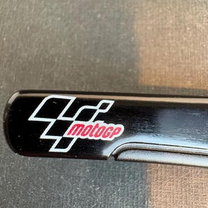 May include: A black rectangular object with the MotoGP logo. The logo features a white and red design with the text "motogp" in red. The object has a curved edge.