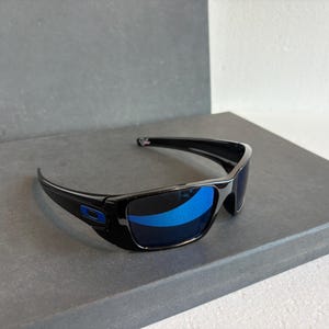 May include: Black sunglasses with blue mirrored lenses. The sunglasses have a rectangular frame with a subtle curve and a blue logo on the side. The lenses reflect the surrounding environment.