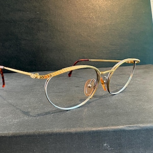Peut inclure: Lunettes dorées de forme œil de chat. Les montures présentent un design délicat avec une finition dorée, complétées par des verres transparents. Les branches ont un motif écaille de tortue aux extrémités. Les lunettes sont posées sur un fond sombre.