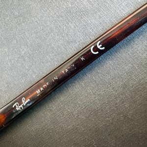 May include: A dark brown tortoiseshell patterned eyeglass temple arm featuring the white printed text Ray-Ban, MADE IN ITALY K, and the CE mark. The arm has a smooth, glossy finish and a metallic hinge component visible at the bottom left corner.