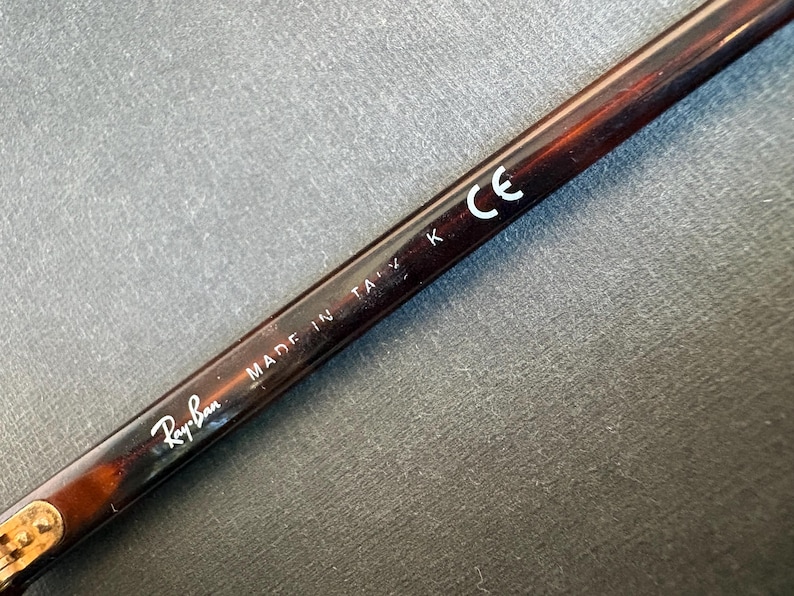 May include: A dark brown tortoiseshell patterned eyeglass temple arm featuring the white printed text Ray-Ban, MADE IN ITALY K, and the CE mark. The arm has a smooth, glossy finish and a metallic hinge component visible at the bottom left corner.