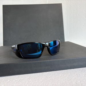 May include: Black sunglasses with blue mirrored lenses. The sunglasses have a rectangular frame and are set against a dark gray background. The lenses reflect the surrounding environment.