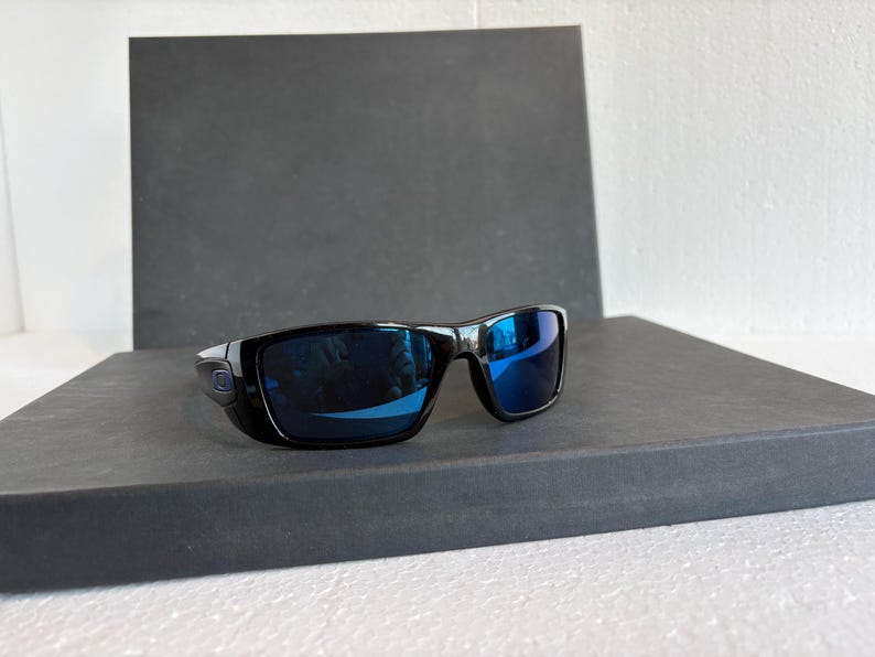 May include: Black sunglasses with blue mirrored lenses. The sunglasses have a rectangular frame and are set against a dark gray background. The lenses reflect the surrounding environment.