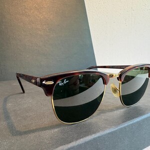 May include: Ray-Ban Clubmaster sunglasses featuring a tortoiseshell browline and temples with gold-tone metal rims and bridge. The dark green lenses include the white Ray-Ban logo on the upper right corner and small silver-tone accents on the frame.