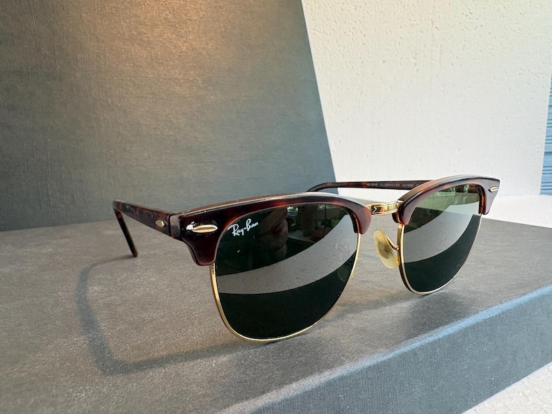 May include: Ray-Ban Clubmaster sunglasses featuring a tortoiseshell browline and temples with gold-tone metal rims and bridge. The dark green lenses include the white Ray-Ban logo on the upper right corner and small silver-tone accents on the frame.