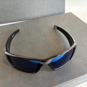 May include: Black sunglasses with blue lenses. The sunglasses have a sleek, modern design with a black frame and a silver accent. The lenses are a vibrant blue color, offering a stylish look.