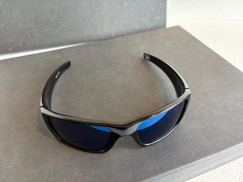 May include: Black sunglasses with blue lenses. The sunglasses have a sleek, modern design with a black frame and a silver accent. The lenses are a vibrant blue color, offering a stylish look.