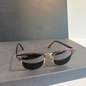 May include: Clubmaster sunglasses featuring a tortoiseshell browline and temples with a gold-tone metal bridge and lower rim. The dark lenses include the Ray-Ban logo on the upper right corner. The temples are thin with a tortoiseshell pattern finish.