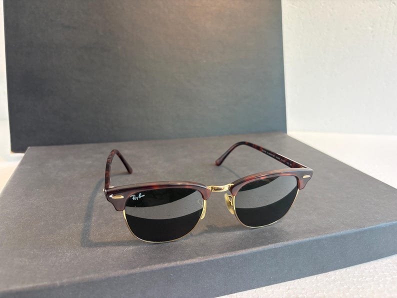 May include: Clubmaster sunglasses featuring a tortoiseshell browline and temples with a gold-tone metal bridge and lower rim. The dark lenses include the Ray-Ban logo on the upper right corner. The temples are thin with a tortoiseshell pattern finish.