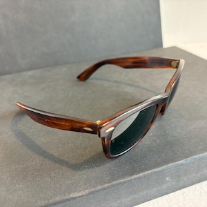 May include: Tortoiseshell sunglasses with dark lenses. The frames are brown and have a classic, rectangular shape with a curved design at the temples. The sunglasses are set against a neutral background.