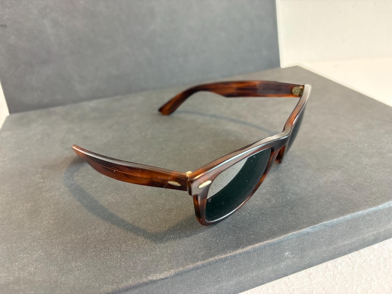 May include: Tortoiseshell sunglasses with dark lenses. The frames are brown and have a classic, rectangular shape with a curved design at the temples. The sunglasses are set against a neutral background.