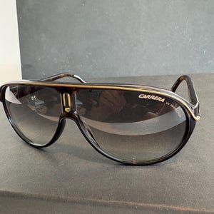 May include: Dark brown Carrera sunglasses with a gradient lens. The sunglasses have a thick frame with gold accents and the Carrera logo on the side. The lenses are a dark grey colour, and the frame has a tortoiseshell pattern.