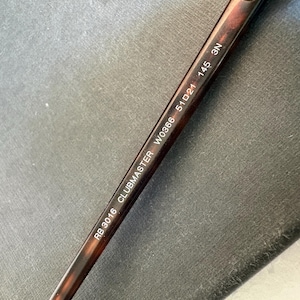 May include: A brown tortoiseshell patterned temple arm of a pair of sunglasses with white printed text that reads RB 3016 CLUBMASTER W0366 51 21 145 3N. The arm is made of a smooth material and features a curved design for resting behind the ear.