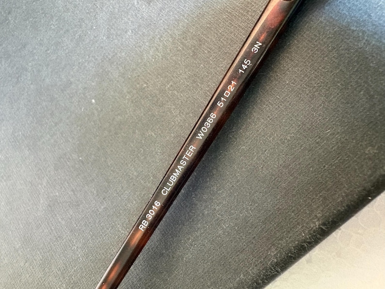 May include: A brown tortoiseshell patterned temple arm of a pair of sunglasses with white printed text that reads RB 3016 CLUBMASTER W0366 51 21 145 3N. The arm is made of a smooth material and features a curved design for resting behind the ear.