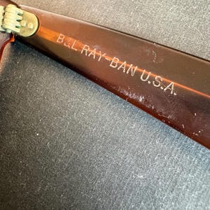 May include: Close-up of the arm of a pair of brown Ray-Ban sunglasses. The text "B&L RAY-BAN U.S.A." is etched in white on the arm. The hinge is visible, with a gold-coloured metal.