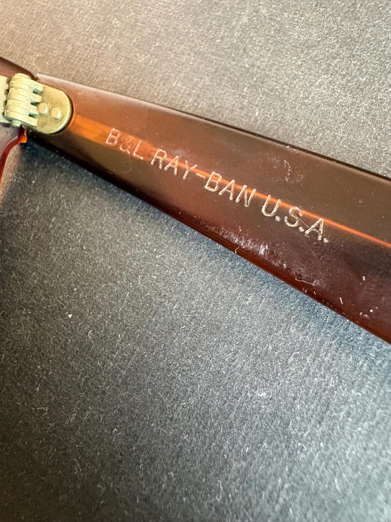 May include: Close-up of the arm of a pair of brown Ray-Ban sunglasses. The text "B&L RAY-BAN U.S.A." is etched in white on the arm. The hinge is visible, with a gold-coloured metal.
