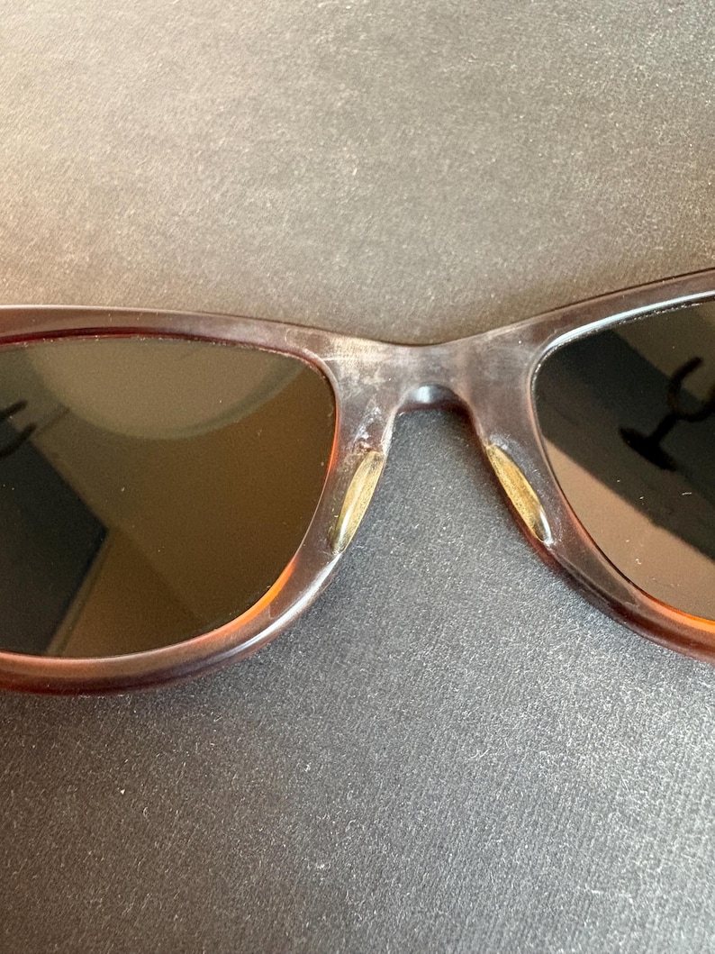 May include: Close-up of brown sunglasses with dark lenses. The frames have a mottled, translucent appearance. The lenses are dark, and the bridge of the glasses is visible.