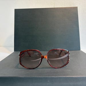 May include: Oversized sunglasses featuring a glossy, dark brown tortoiseshell frame with a rounded, slightly angular shape. The lenses are tinted in a gradient brown hue, transitioning from a darker shade at the top to a lighter shade at the bottom.